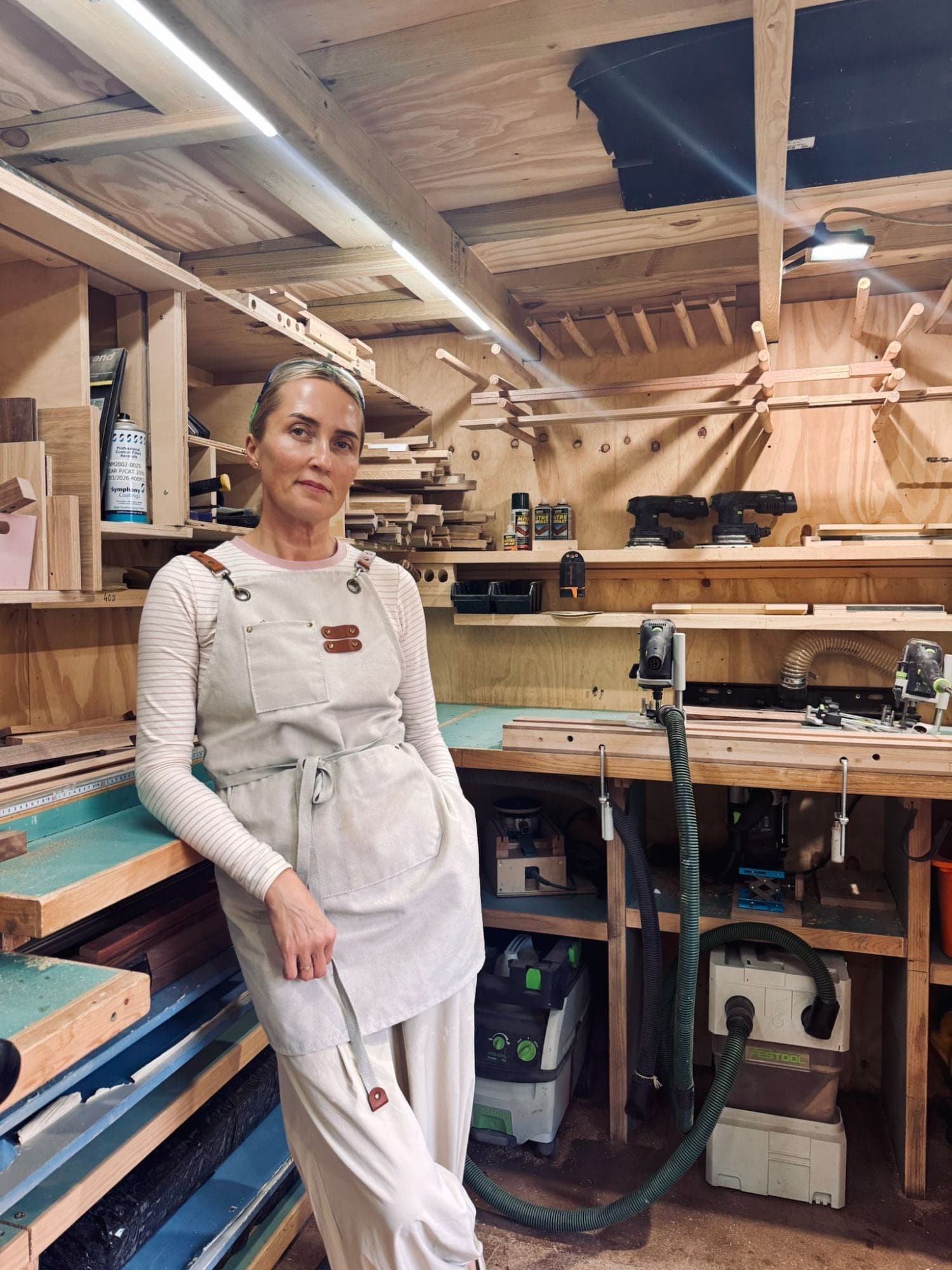 Kat leaning on a workbench in the workshop, in front of the timber racks and tools