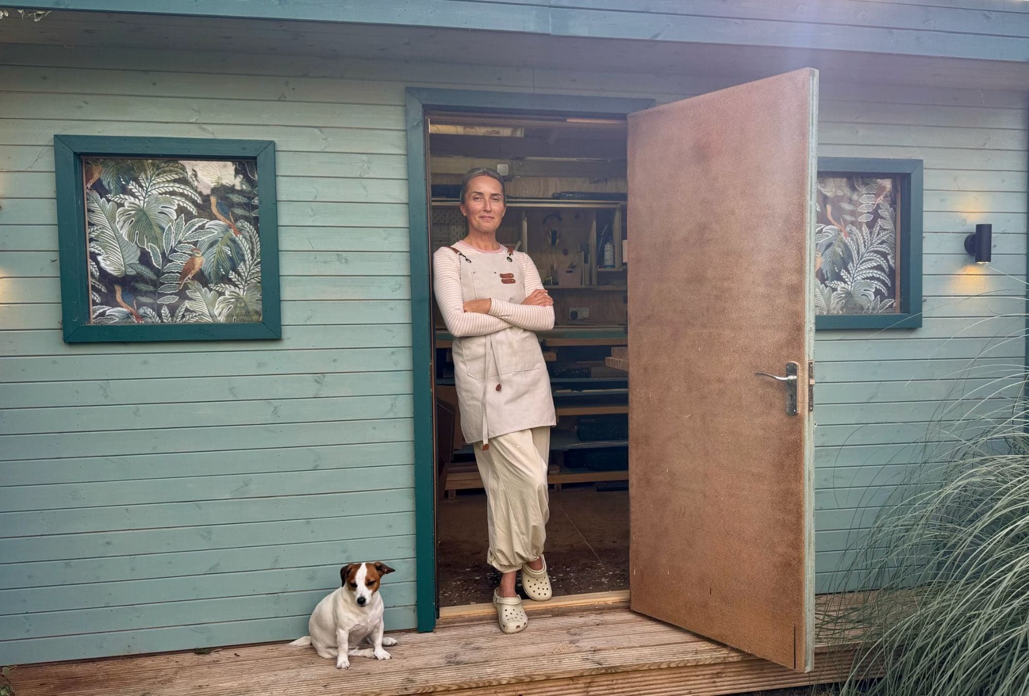 Kat leaning in the doorway of the WoodlandByKat workshop, with the dog sitting on the decking